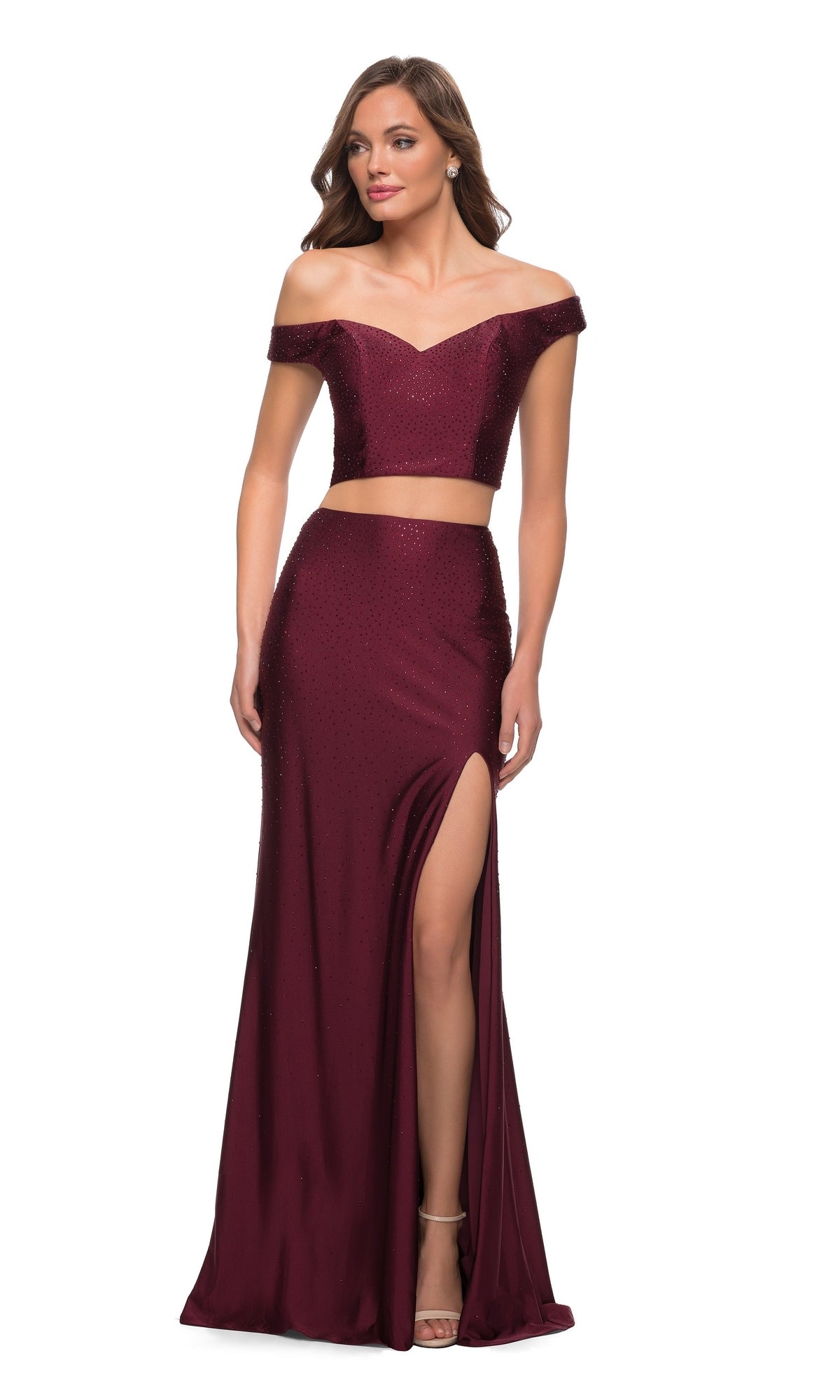 A woman wears the La Femme 29951, an off-the-shoulder two-piece prom dress in burgundy featuring a thigh-high slit and subtle sparkle, paired with strappy heels.